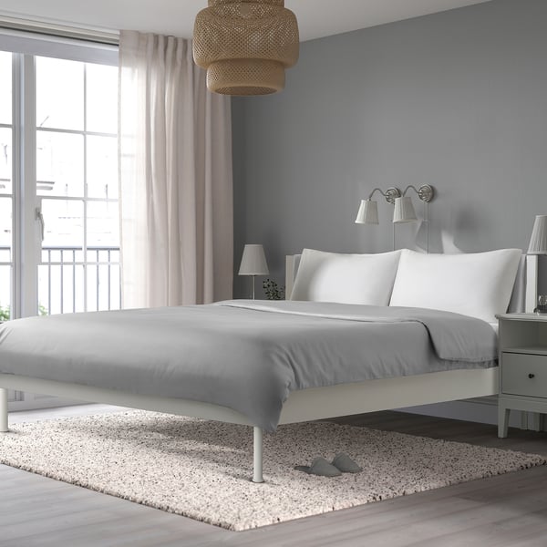 A modern bedroom with a white BRUKSVARA bed on a speckled rug. The bed has a cushioned headboard and ample space underneath for storage.