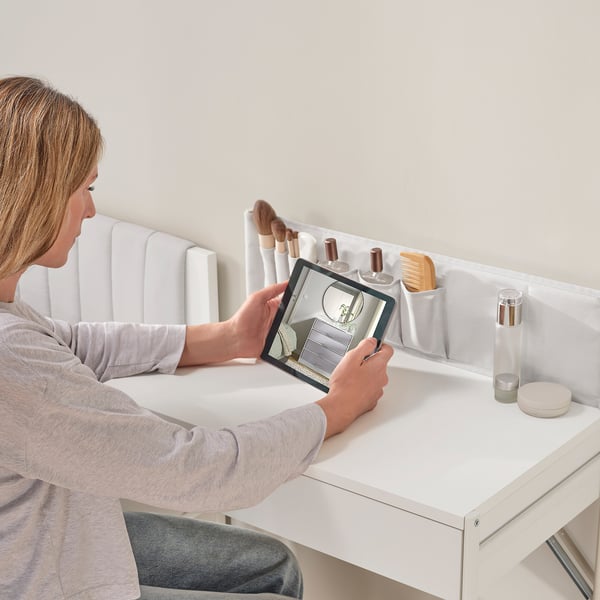 A person sits at a dressing table, using a tablet. The table has a drawer, fabric pockets with brushes, and beauty items.