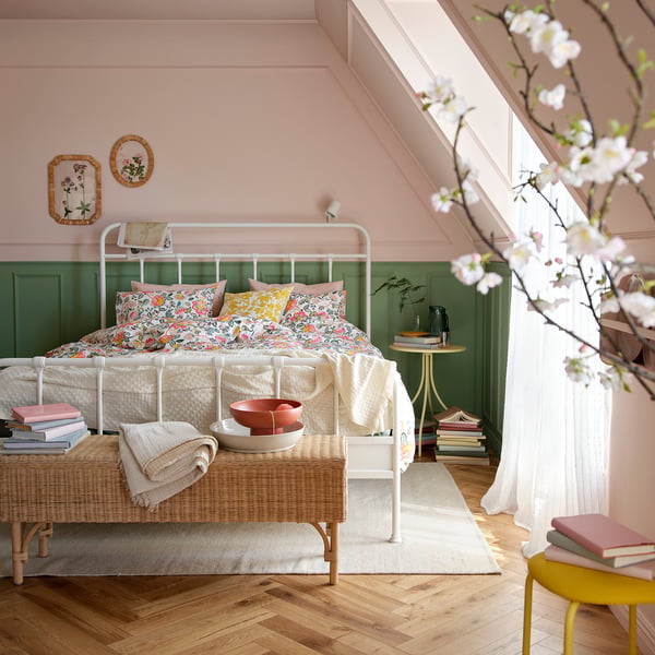 Pastel duvet bed, green wainscoting, pink walls, wooden floors, wicker bench with books.