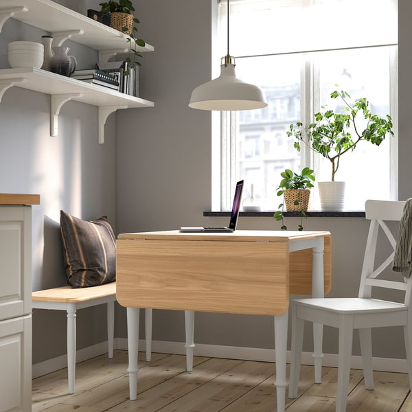 Drop-leaf table by window in minimalist kitchen, fits under with plant and laptop. Shelves, chair with cushion nearby.