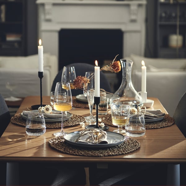 Wooden drop-leaf table set for two. Dark plates, glasses, candles, and dried flowers. Cosy room with sofa and tv.