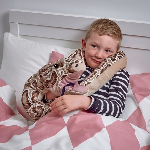 A child hugs a large plush python toy on a bed, the toy a pattern and a pocket in its head.
