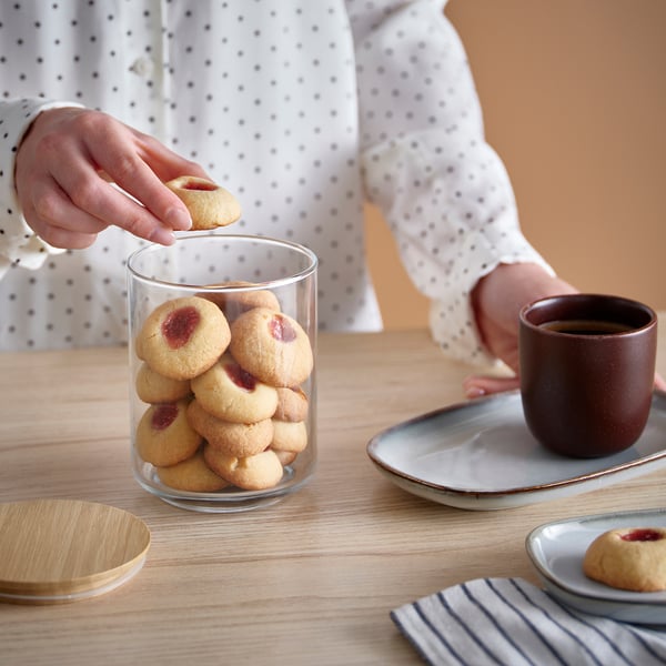 A person reaches for a cookie from a glass jar filled with thumbprint cookies on a wooden table, accompanied by a cup of coffee.