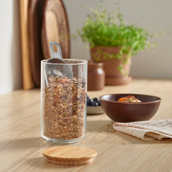 Glass jar labelled EKLATANT holds granola, with bamboo lid and metal scoop. Nearby, wooden table has a plant and spoons.