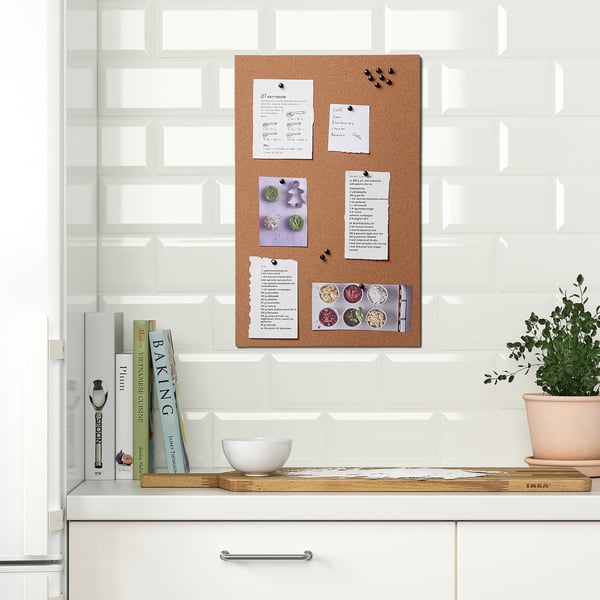 Kitchen wall with FLÖNSA cork memo board displaying notes and photos, surrounded by cookbooks and a potted plant, serving as an organisational hub.