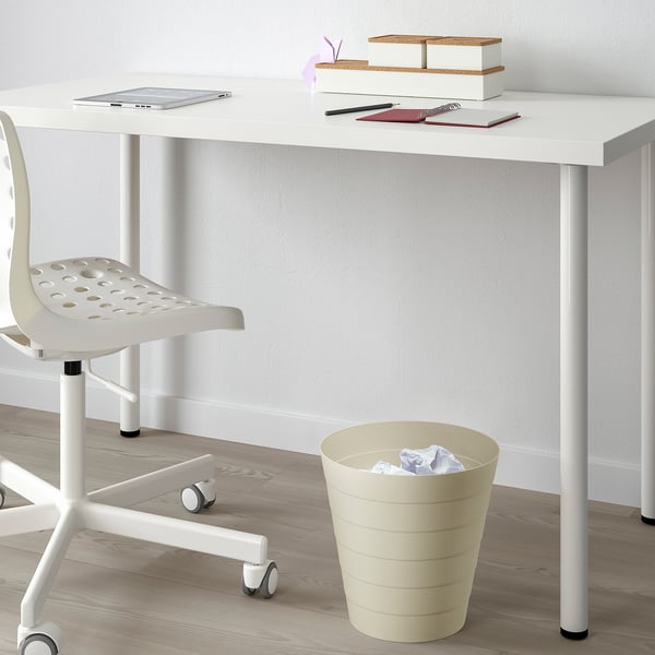 A minimalist workspace with a white desk, a white chair, and a beige waste bin holding paper. The desk has a laptop, notebooks, and storage boxes.