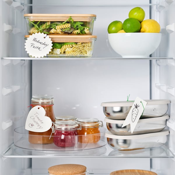 Open refrigerator with organised shelves, showing labelled meal containers, jars of preserves, and a bowl of citrus fruits.