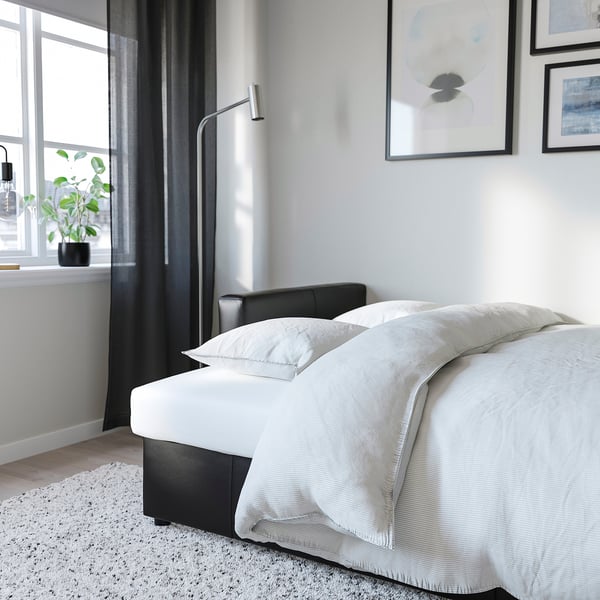 Bed with storage underneath, light grey bedding, metal lamp, black curtain, and potted plant near window.