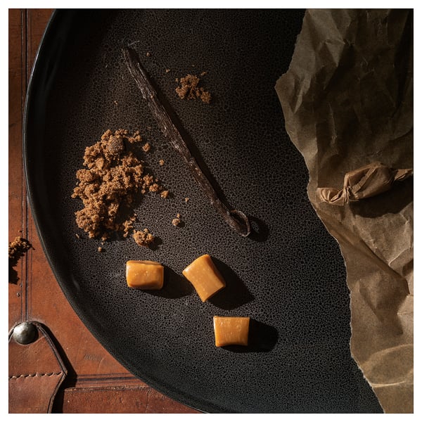 Black plate with scattered caramel candies, a vanilla bean, and crumbs. Rustic, brown paper backdrop.