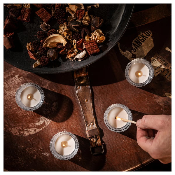 Four lit candles in silver holders on a wooden table, surrounded by dried plant material in a black bowl; person holding a matchstick.