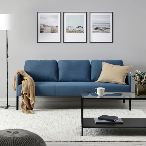 Modern living room with blue GLOSTAD sofa, grey pillows, black coffee table, white rug, and coastal art.