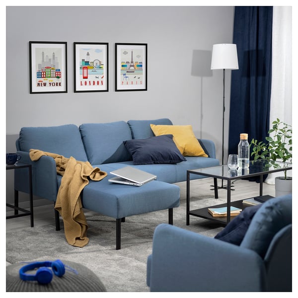 Modern living room with blue GLOSTAD sofa, yellow pillows, city prints on wall, floor lamp, coffee table with plants, laptop, glass, and headphones.