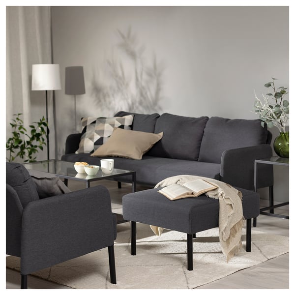 Modern living room with a grey GLOSTAD sofa, footstool, and armchair. Decor includes a glass coffee table, floor lamp, and plants.
