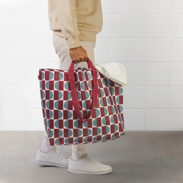 Person holding large red and blue patterned tote bag with white trainers.