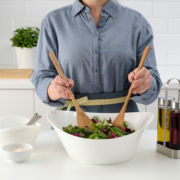 Person in blue shirt tossing salad with wooden tongs in large white bowl on kitchen counter.