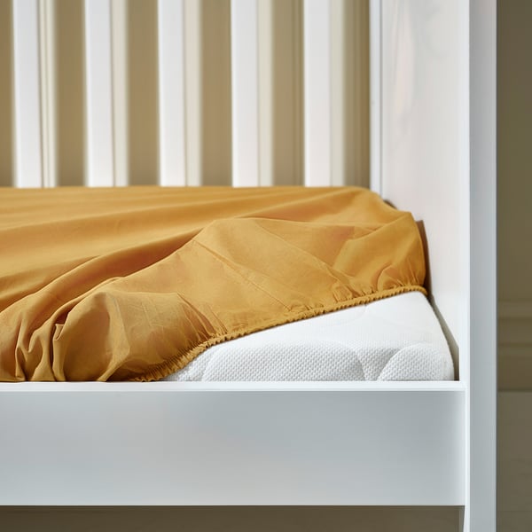 White crib with yellow fitted sheet, showing safety elastic edges for secure fit on mattress.