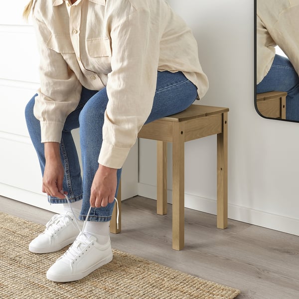 Person tying shoelaces while sitting on a wooden stool. The stool has a cut-out handle for easy carrying.