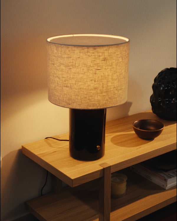 Modern HALGATT table lamp with beige linen shade, brass knob, and dark base on shelf, emitting warm light.