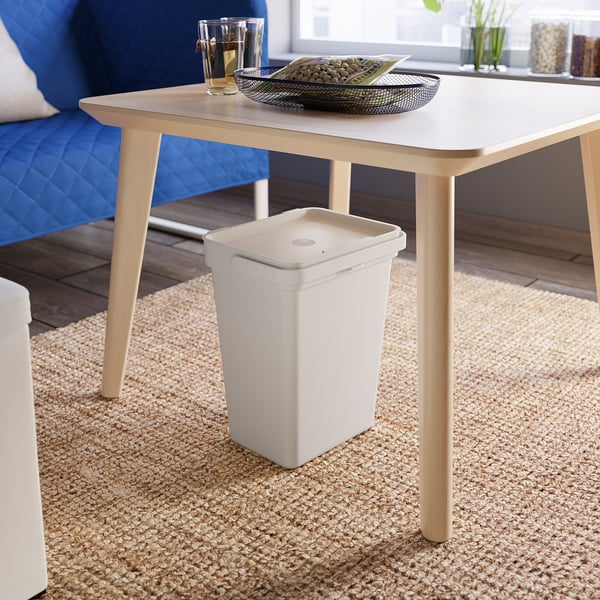 A white HÅLLBAR bin under a light wooden table. The bin has a lid and handle, placed on a jute rug.