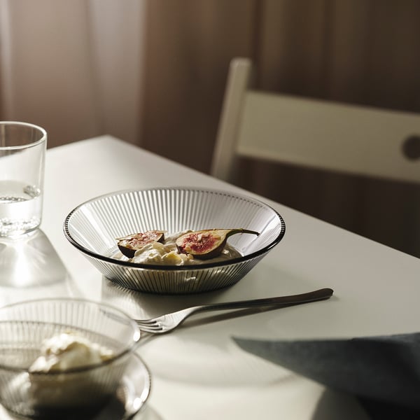 A white bowl with black stripes holds sliced fruit and yoghurt, placed on a white tablecloth with a glass of water and a fork.