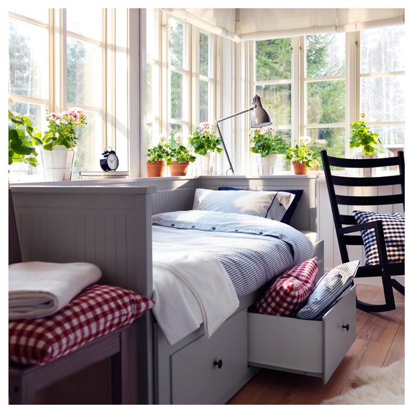 Cosy bedroom with HEMNES grey day-bed, red-checkered pillows, white storage, black chair, plants, large windows, wooden floor, soft lighting.