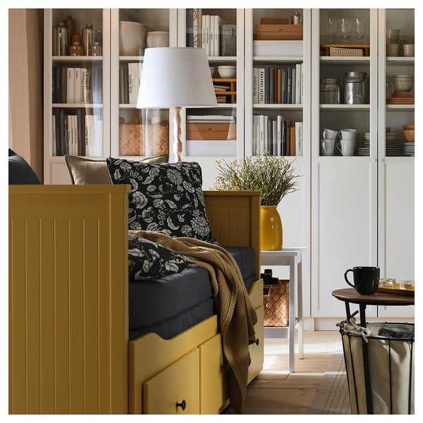 Bright yellow HEMNES day-bed with storage drawers and shelves, with a black and white floral pillow, a side lamp, and a vase with greenery.