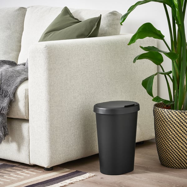 Small HÖLASS black bin sits beside a light grey sofa, near a large house plant. Its cylindrical, made of polypropylene plastic, and has a lid.
