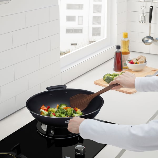 Cooking vegetables in wok. One hand holds wok, other adds broccoli. Kitchen counter has utensils and ingredients.