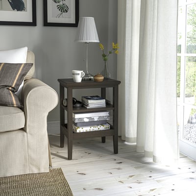 Wooden side table IDANÄS with shelves, beside armchair, grey & natural wood, near window with curtains.