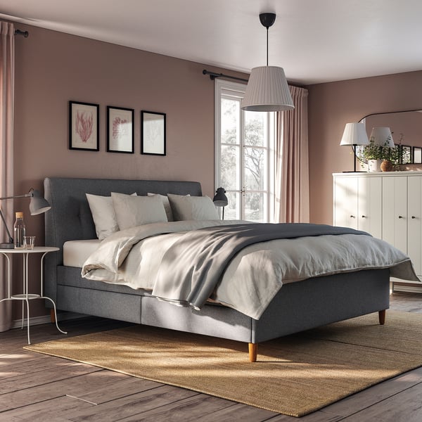 Modern bedroom with IDANÄS grey upholstered bed, white curtains, and pendant light. Neutral decor with rug, plants, and wall art.