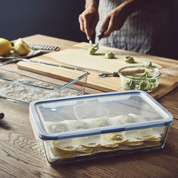 A person prepares food on a wooden board. A glass food container with a lockable lid stores layered food items.