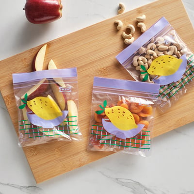 Three purple IKEA ISTAD bags hold nuts, dried fruits, and apple slices, sealed with lemon stickers, on a wooden cutting board.