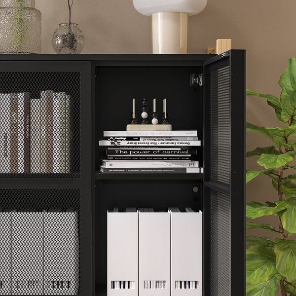 Black IVAR storage unit with open door revealing books and records. Mesh doors on either side, placed against wall with decor items.