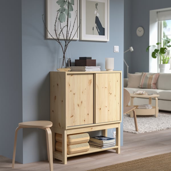 IVAR light wood cabinet in living room, topped with books and candle, tall plant next to it, beneath framed art and clock.