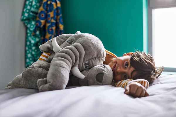 A child hugs a large grey elephant cuddly toy on a bed. The toy is soft with white tusks and floppy ears.