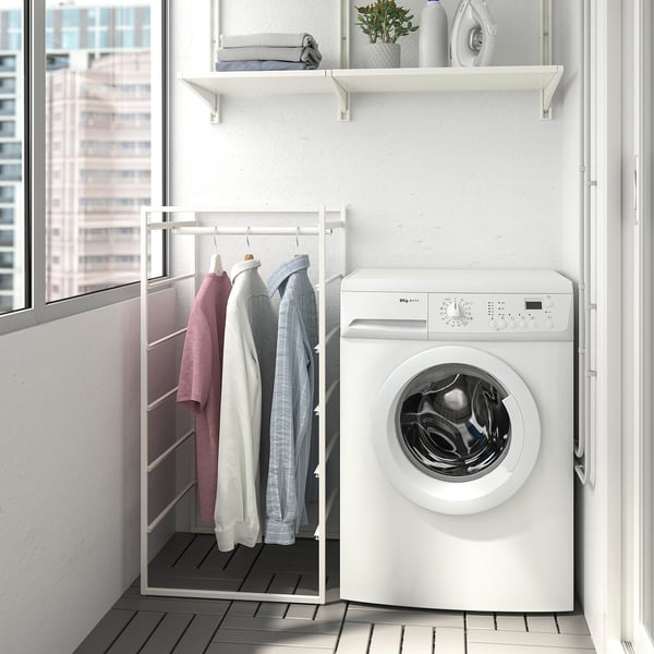 Small white JONAXEL clothes rack between washer & window, holding pink, white, & blue shirts, fitting neatly in compact room.