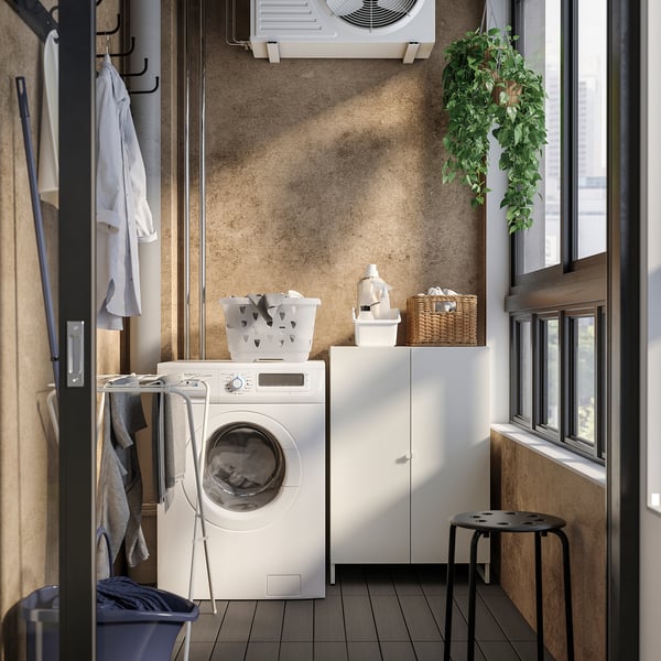 Small laundry room with white JOSTEIN storage. Washer, hanging shirts, baskets, and a hanging plant. Floor dark, wall beige.