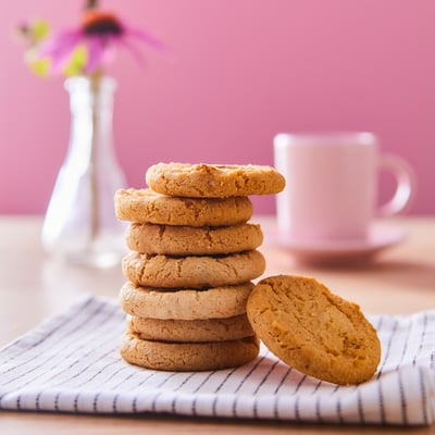 A stack of golden-brown KAFFEREP cookies on a striped cloth, with a white mug and vase with flowers in the background.