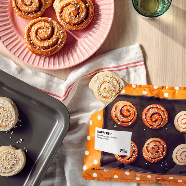Image shows cinnamon buns on trays, some baked and others in packaging labelled KAFFEREP from IKEA, with a glass of liquid on a wooden table.