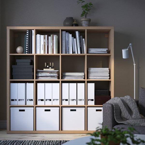 A KALLAX shelf in a room, filled with books, decor, and boxes. To the right, a grey chair and floor lamp with a patterned rug underneath.
