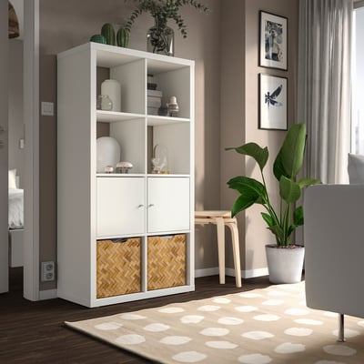 White KALLAX shelving with mix of open shelves, cabinets, and baskets, positioned against a wall with plants and artwork, larger than nearby chair.