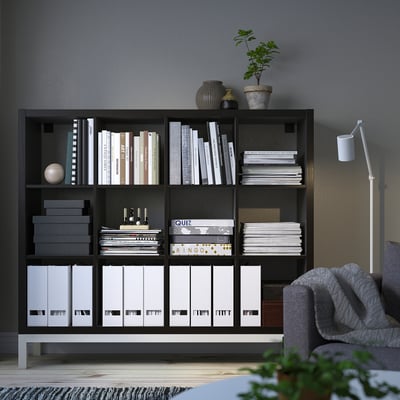 Black KALLAX bookshelf against grey wall. Holds books and decor. Tall, rectangular, airy design with underframe. Grey sofa and plant nearby.
