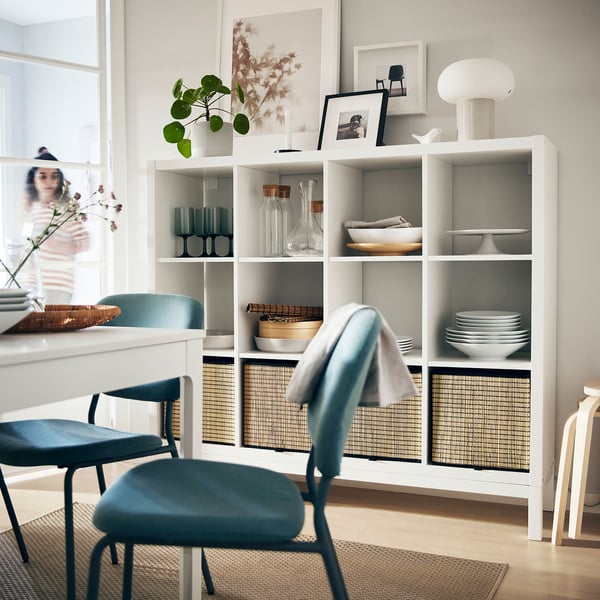 Modern dining area with white KALLAX shelf, blue cushioned chairs and various decorative items including glasses, plant, dishes, and framed photos.