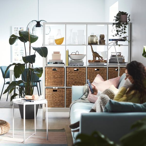 Living room with blue sofa, coffee table, KALLAX shelves, books, decor, person on smartphone.