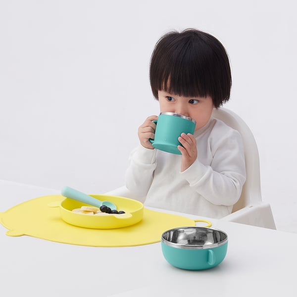 Child drinking from stable turquoise mug with large handle, sitting at white table with yellow silicone accessories.