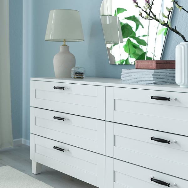 White dresser with black handles, lamp, books, and mirror with plant reflection.