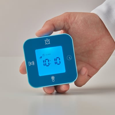 Hand holding blue KLOCKIS clock. Displays 10:10, alarm icon shown. Simple, multifunctional design.