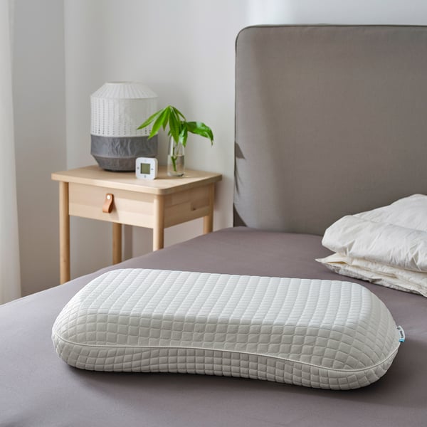 Grey ergonomic pillow on bed, memory foam & gel layer. Beside: lamp, clock, plant.