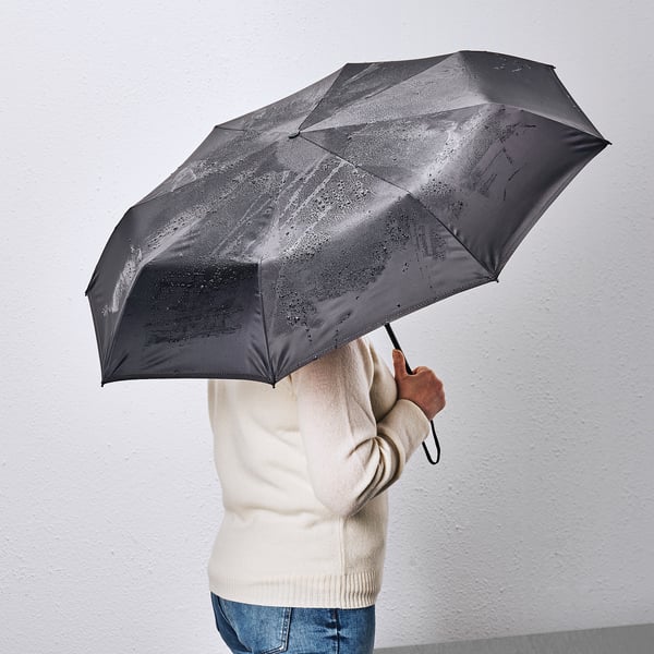 A person holding a black KNALLA umbrella, which is foldable and provides protection from heavy rain. The handle has a strap for easy carrying.