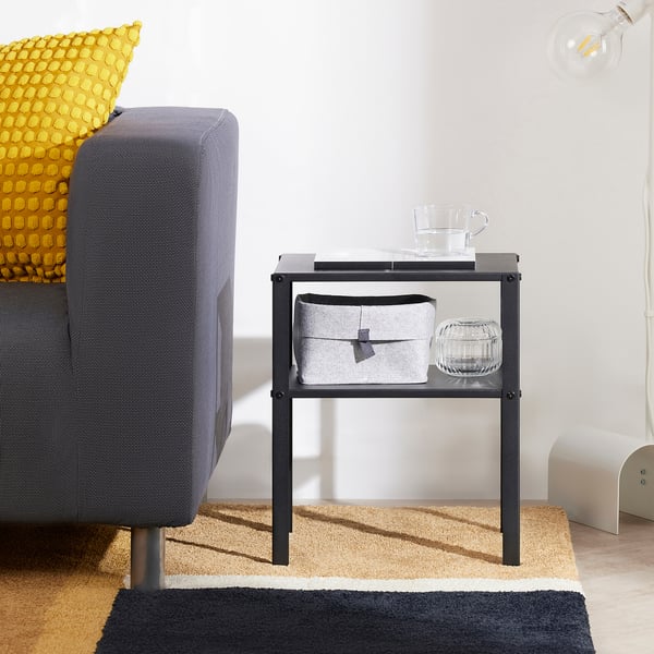 Small black KNARREVIK bedside table next to a grey chair. Holds a glass cup, book, and storage box, ideal for small spaces.